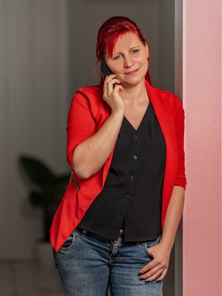 Buche deine Transformation Frau mit rotem Blazer und schwarzem Oberteil, die am Telefon lächelt.