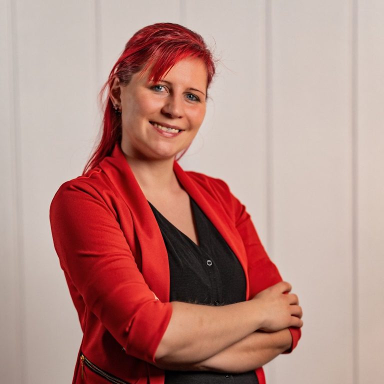 Beginne jetzt deine Transformation Frau mit rotem Blazer und roten Haaren, lächelnd mit verschränkten Armen.