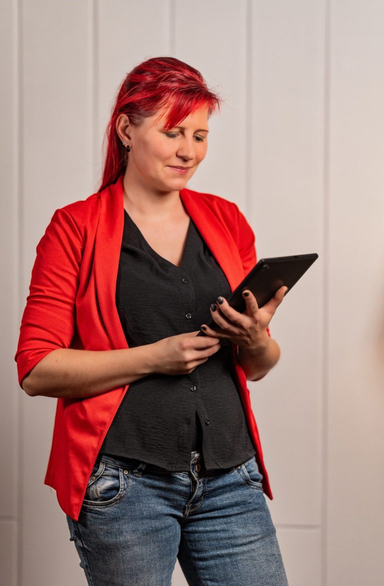 Kostenloses Gespräch buchen Frau mit roten Haaren und rotem Blazer, die ein Tablet in der Hand hält.