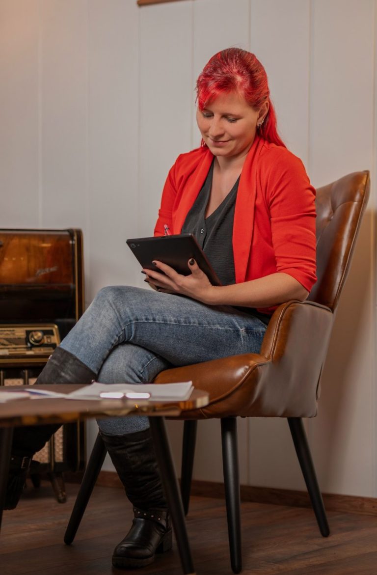 Kostenloses Gespräch buchen Frau mit roten Haaren und rotem Blazer, die ein Tablet in der Hand hält.